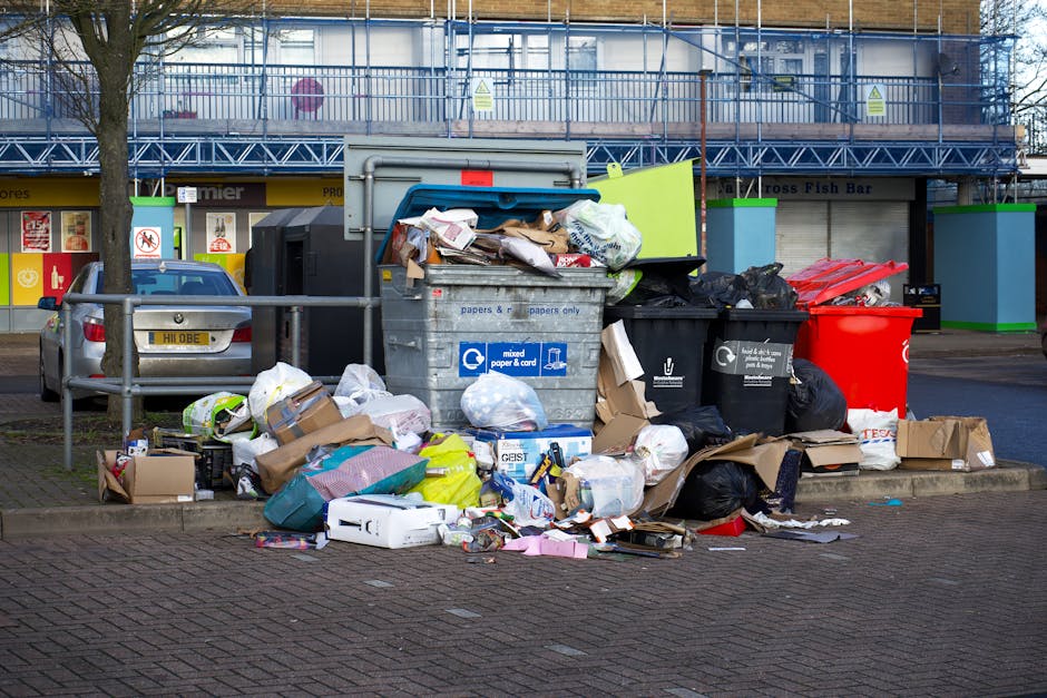A narrow urban alleyway filled with a large, black garbage bag in the foreground, placed on a pavement surface that appears stained and littered with small debris. Behind the bag, there are stacks of 